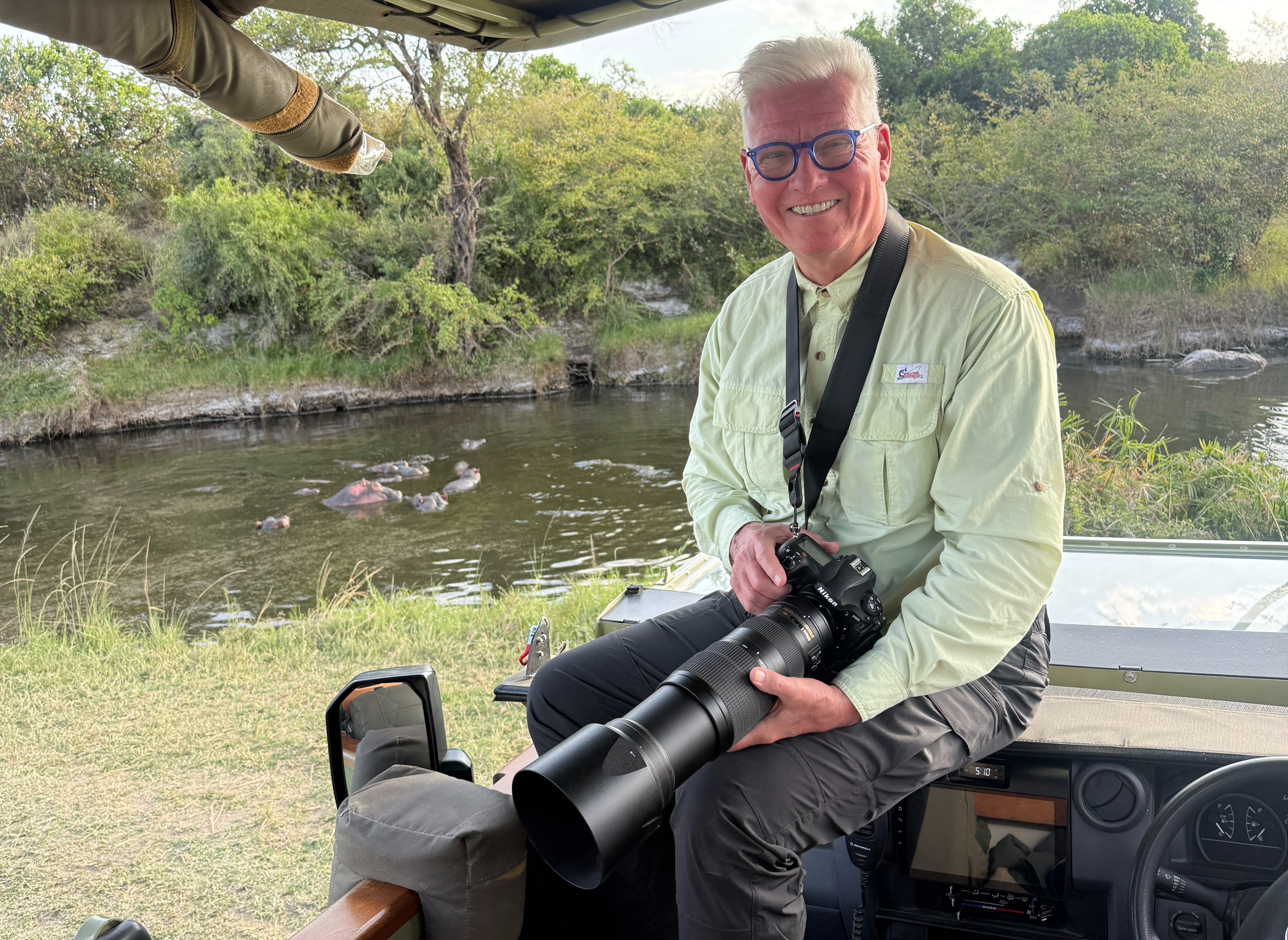 Harry Martin with a telephoto camera on a safari vehicle, hippos in the river behind him